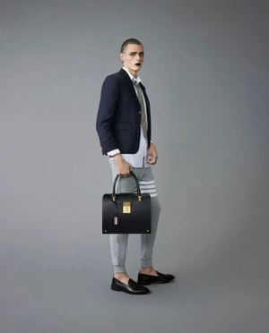 Model in navy blazer, white shirt, and grey sweatpants holding a black leather handbag against a grey backdrop