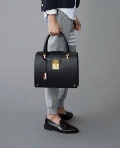 Close-up of a model holding a structured black leather handbag, wearing grey sweatpants and loafers against a grey background.