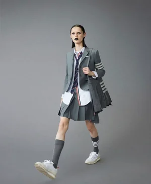 Model in grey blazer, white shirt, skirt, tie, knee-high socks, and sneakers carrying a grey bag against a plain grey backdrop.