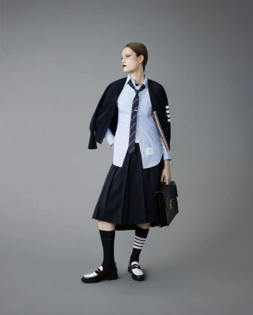 Model wearing a navy pleated skirt, light blue button-down shirt, tie, and dark cardigan carrying a black briefcase.