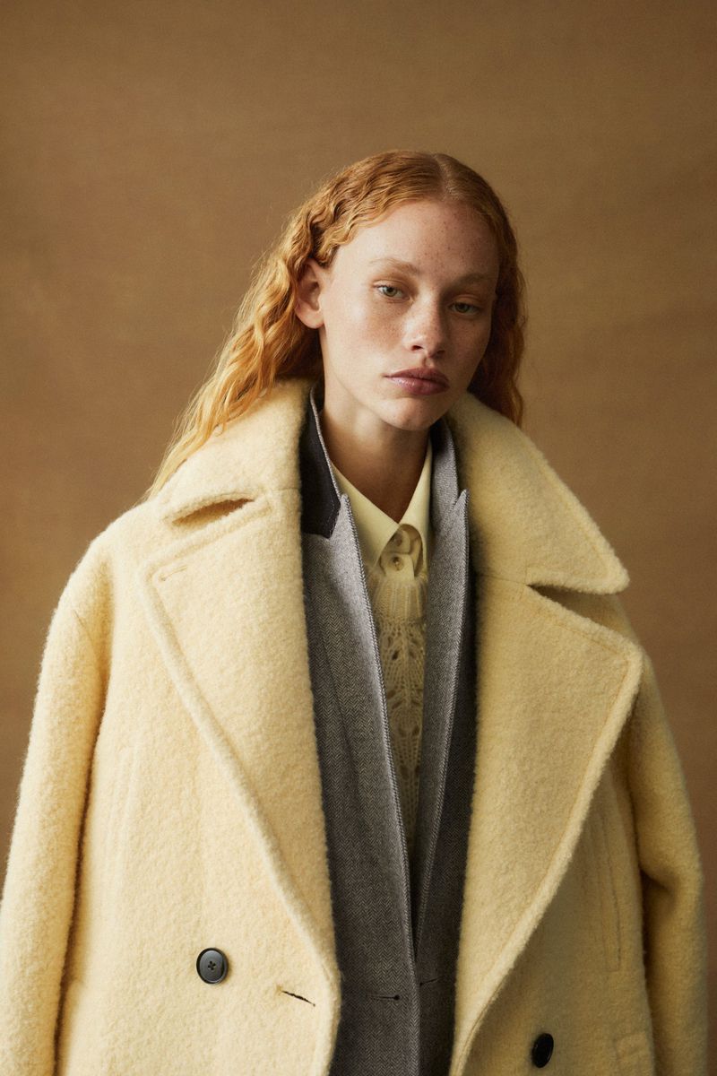 A fair-skinned woman with red hair wearing a cream-colored coat and layers of knits against a neutral brown backdrop in a fashion portrait.