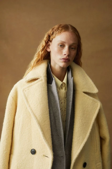 A fair-skinned woman with red hair wearing a cream-colored coat and layers of knits against a neutral brown backdrop in a fashion portrait.