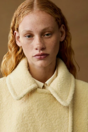 Close-up portrait of a fair-skinned woman with red hair, wearing a yellow, textured coat, posing in an indoor studio setting.