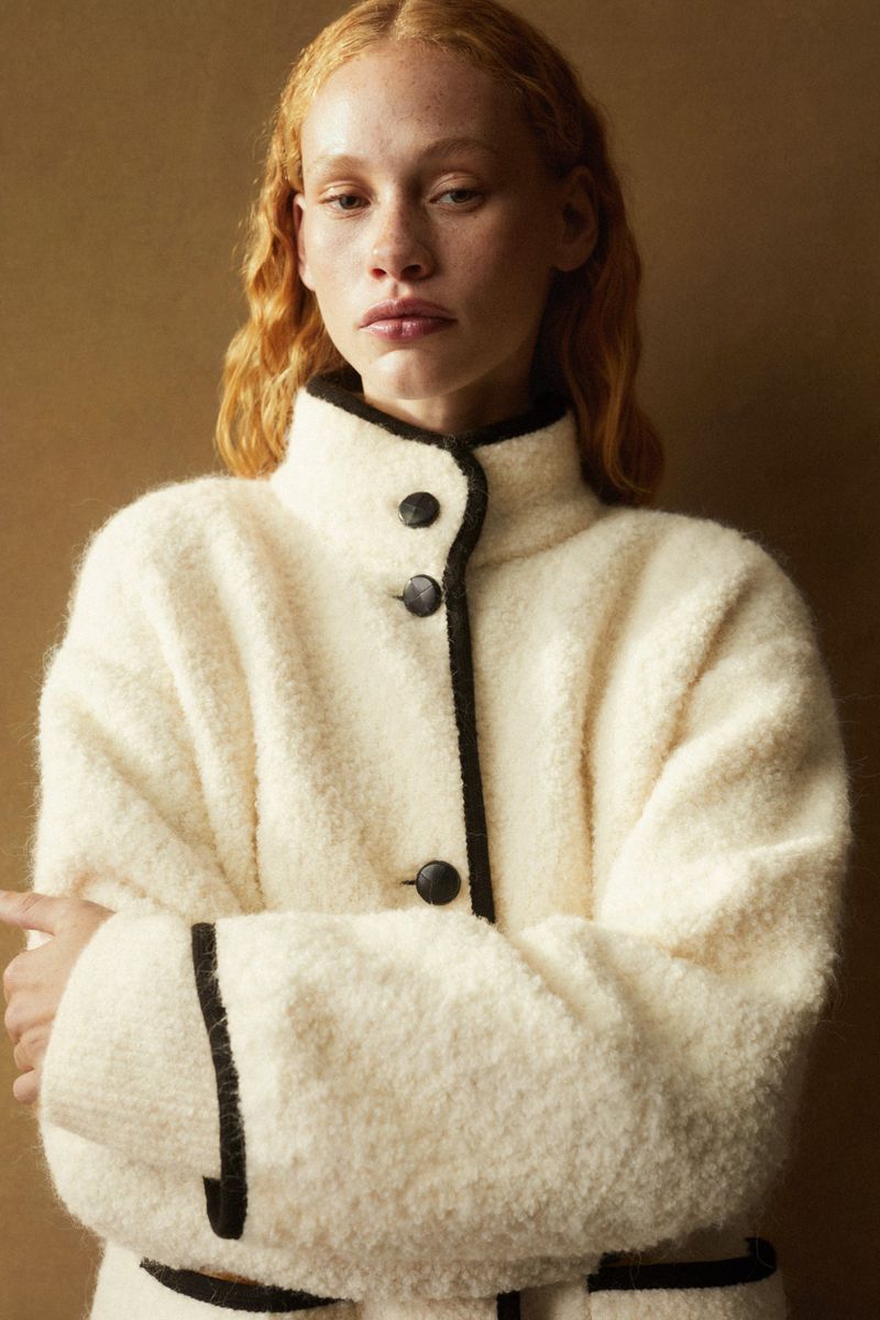 A portrait shot showcases a red-haired woman in a fuzzy, cream-colored coat with black trim, complemented by a simple brown background.