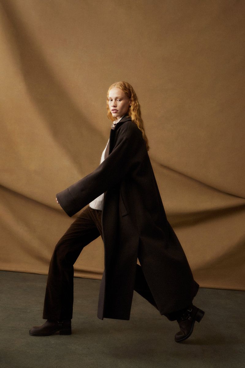 A woman with red hair poses in a full-length dark coat and brown pants against a mottled brown background in a fashion studio setting.
