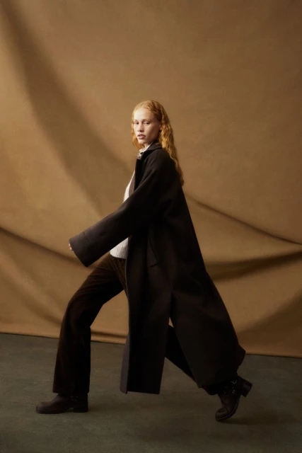A woman with red hair poses in a full-length dark coat and brown pants against a mottled brown background in a fashion studio setting.