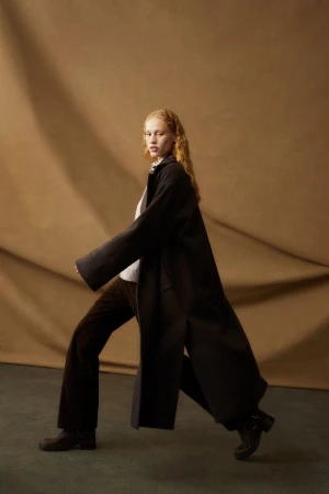 A woman with red hair poses in a full-length dark coat and brown pants against a mottled brown background in a fashion studio setting.