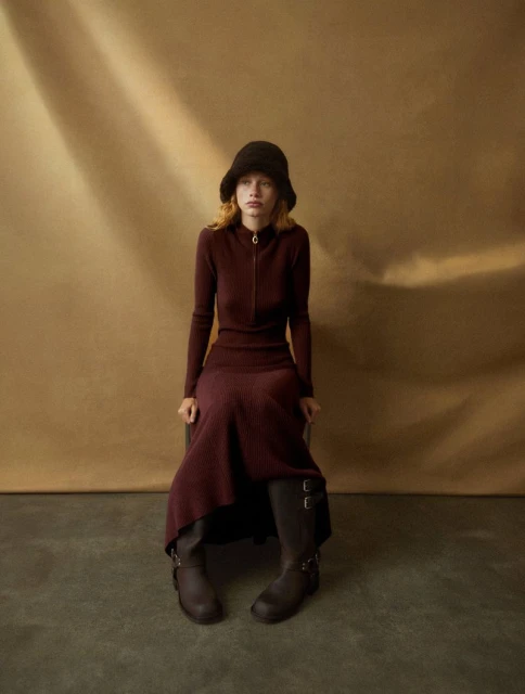 A woman in a brown dress and hat sits indoors with natural lighting, showcasing a vintage-inspired fashion look with a neutral backdrop.