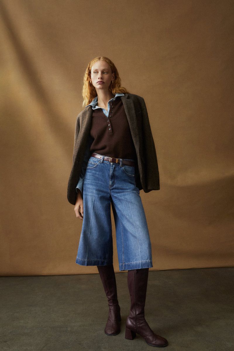 A red-haired woman poses in a brown shirt, blue denim culottes, and knee-high boots against a tan backdrop in a fashion shot.