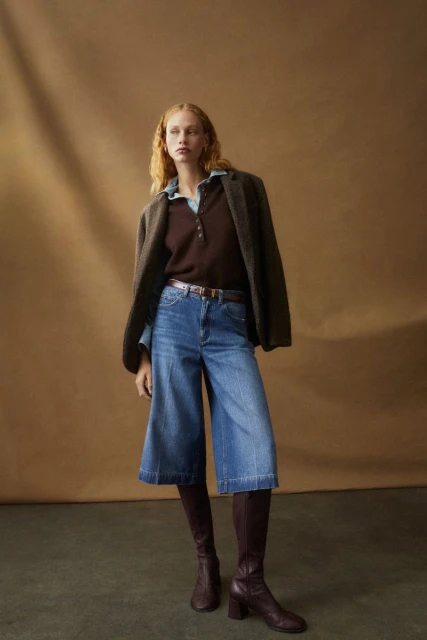 A red-haired woman poses in a brown shirt, blue denim culottes, and knee-high boots against a tan backdrop in a fashion shot.