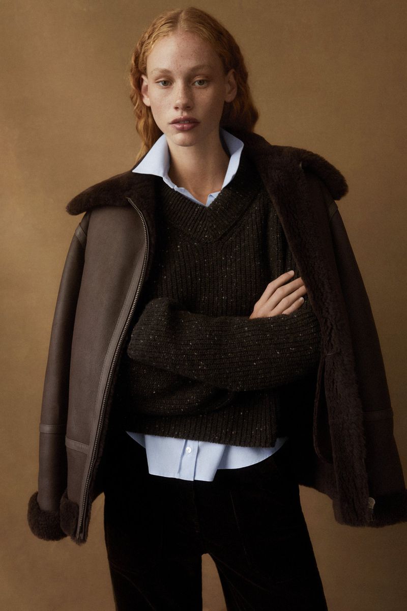 Woman in brown aviator jacket and dark green sweater poses indoors against a neutral backdrop in a fashion portrait. 