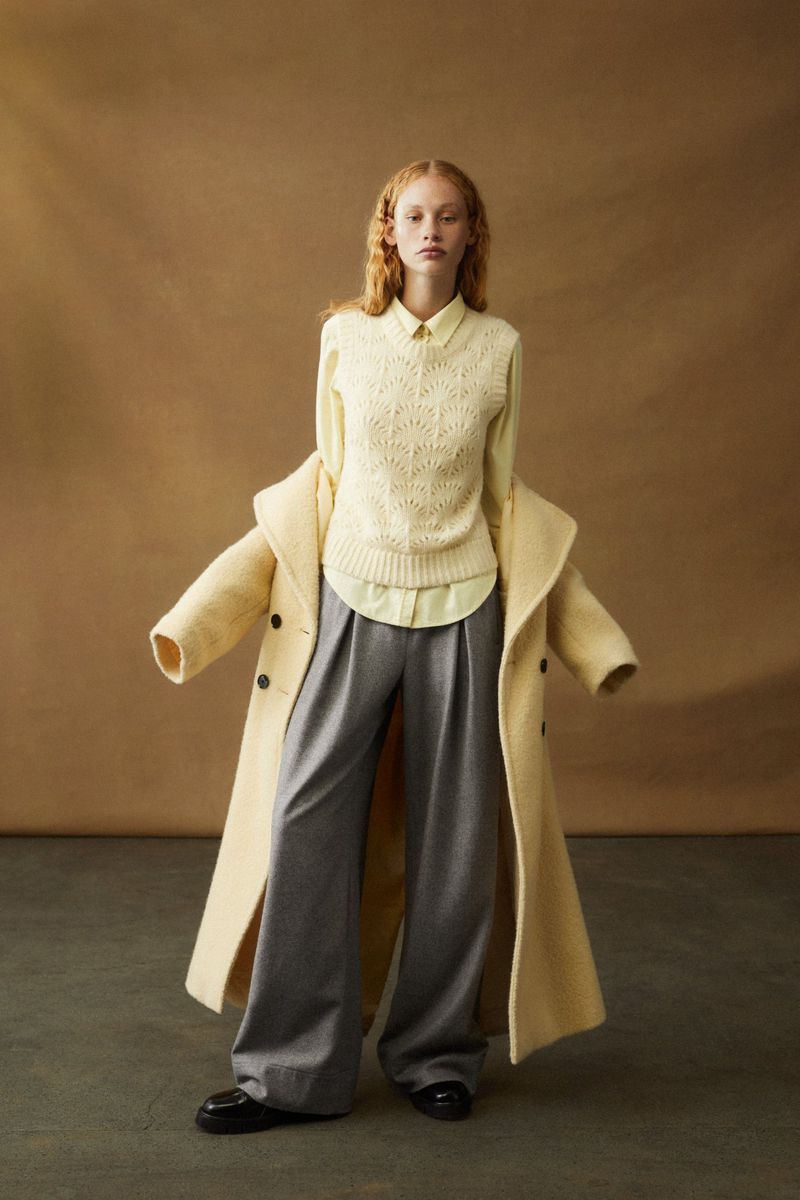 A woman models a yellow sweater vest, collared shirt, and wide-leg grey pants, posing with a long yellow coat in a studio setting.