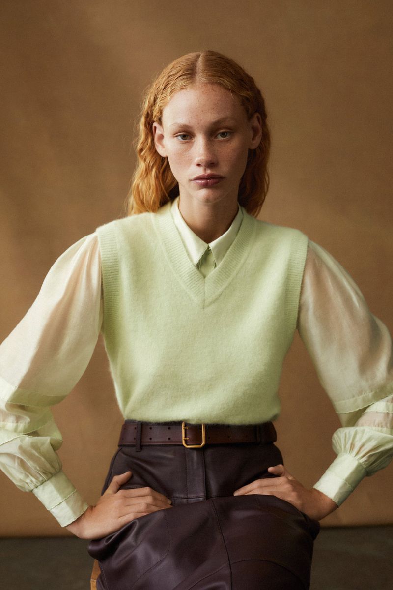 A woman with red hair poses indoors wearing a light green sweater vest, a white collared shirt, brown belt, and dark brown skirt.