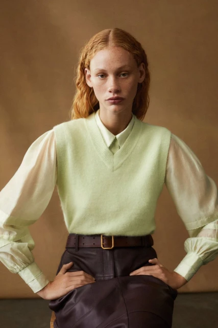 A woman with red hair poses indoors wearing a light green sweater vest, a white collared shirt, brown belt, and dark brown skirt.