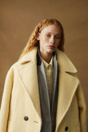 Model with red hair wearing a pale yellow textured coat layered over a grey jacket against a neutral brown backdrop