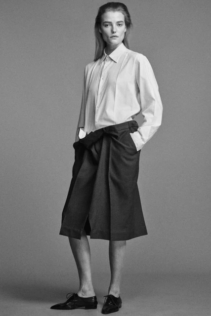 Full shot of a fair-skinned adult woman with long blond hair, wearing a white button-down shirt, dark skirt, and black dress shoes, studio lit.