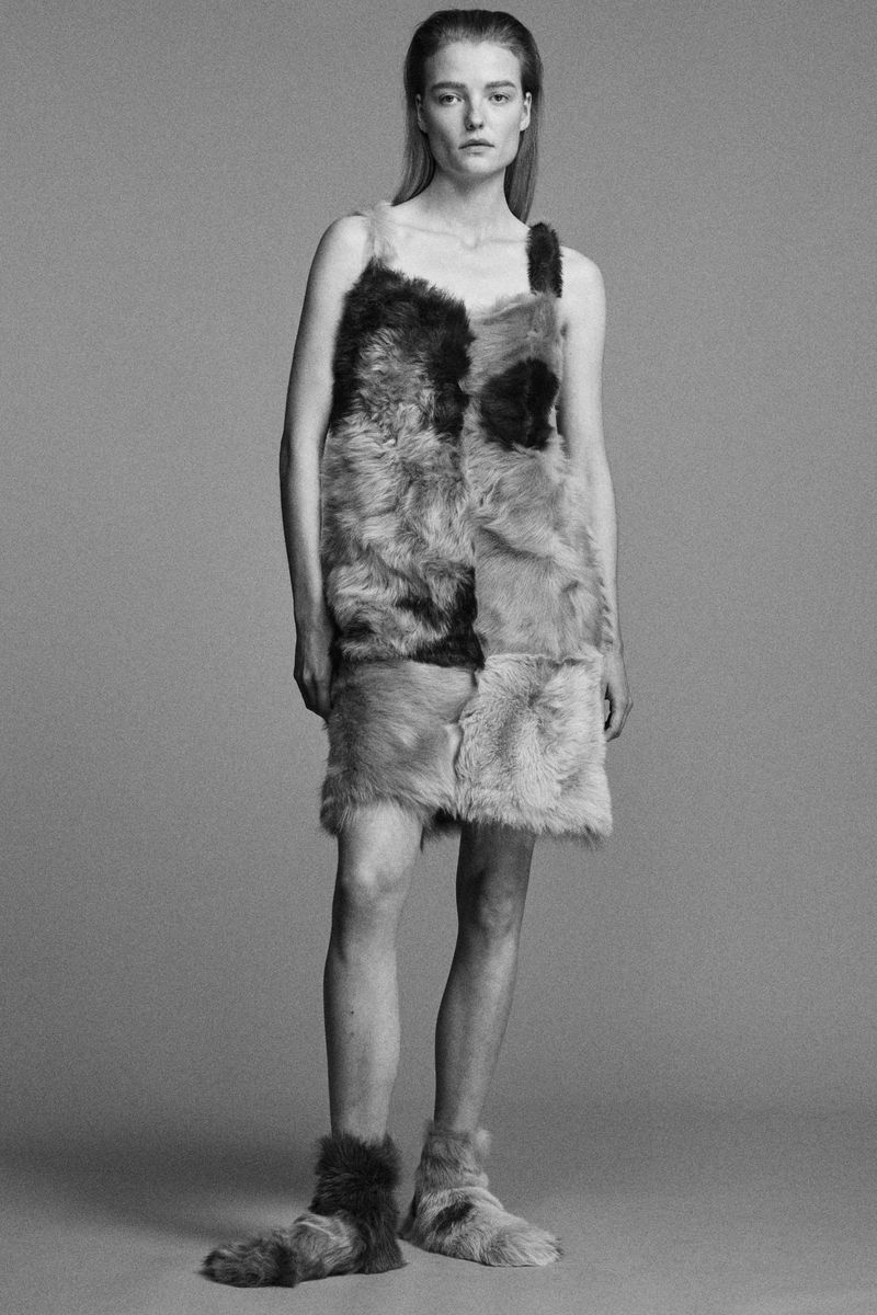 Full shot of a fair-skinned female model posing in a fur patchwork dress and matching furry shoes against a neutral gray backdrop.