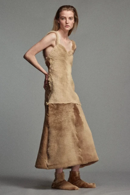 A woman in a tan fuzzy dress and shoes stands in a studio, posing with her hand on her hip, looking forward in a fashion shot.