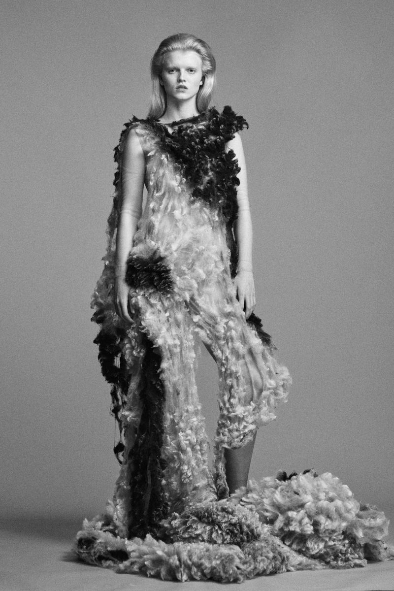 Fashion model in an ornate, textured dress posing in a studio. The high fashion garment features light and dark decorative elements.