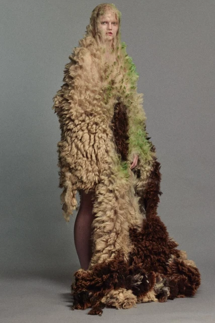 Fashion shot of a slender woman wearing a textured, earth-toned oversized coat or cape posing against a neutral backdrop in a studio.