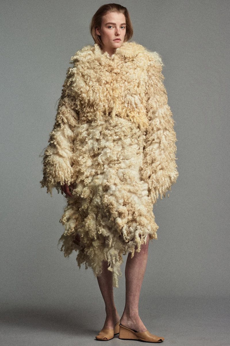 Fashion editorial shot of a model wearing a long, textured beige coat and neutral-toned wedge shoes against a grey studio backdrop.
