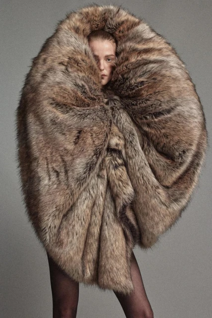 Adult female model in a large, oversized fur coat, standing indoors in a photo studio against a grey backdrop, fashion editorial shot.