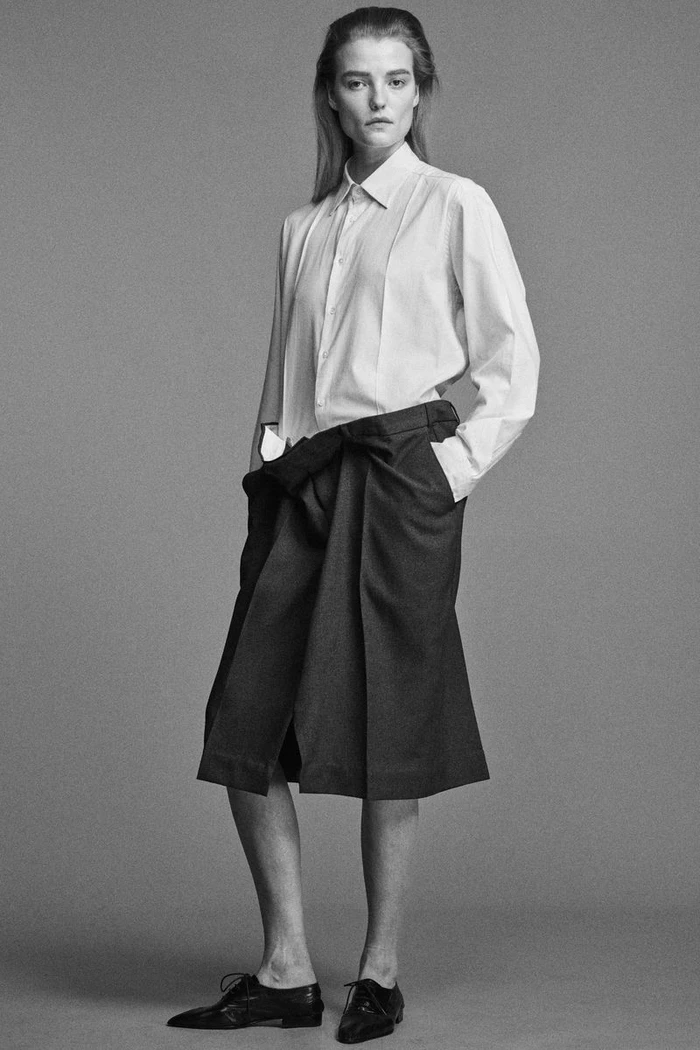 Model in a white shirt and pleated dark skirt standing against a plain grey studio backdrop