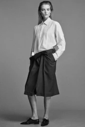 Model in a white shirt and pleated dark skirt standing against a plain grey studio backdrop