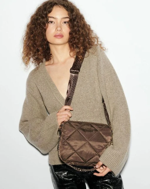 Fashionable woman posing with a quilted crossbody bag in studio setting, wearing a cozy V-neck sweater and shiny black pants.
