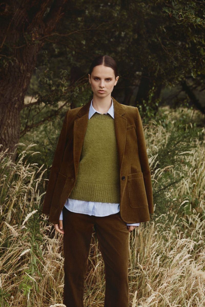 Adult woman in a blazer, sweater, and pants poses outdoors in a grassy field with a wooded background, looking directly at the camera.