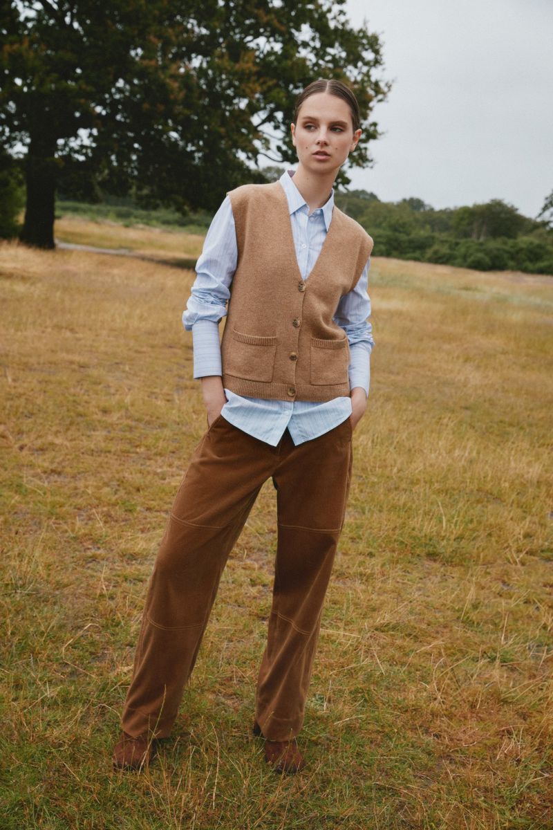 Model standing outdoors on dry grassland wearing a light blue shirt, camel knit vest, and brown trousers.