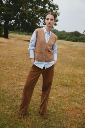 Model standing outdoors on dry grassland wearing a light blue shirt, camel knit vest, and brown trousers.