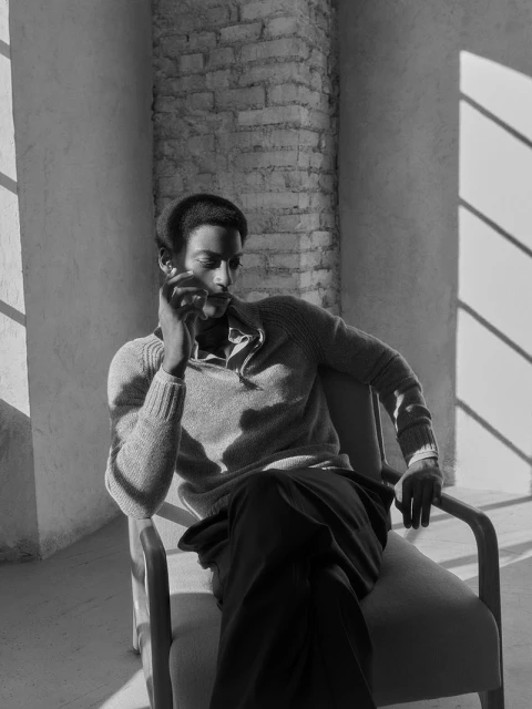 Black and white shot of a Black man in a sweater sitting in a chair near a brick column with natural light casting shadows.