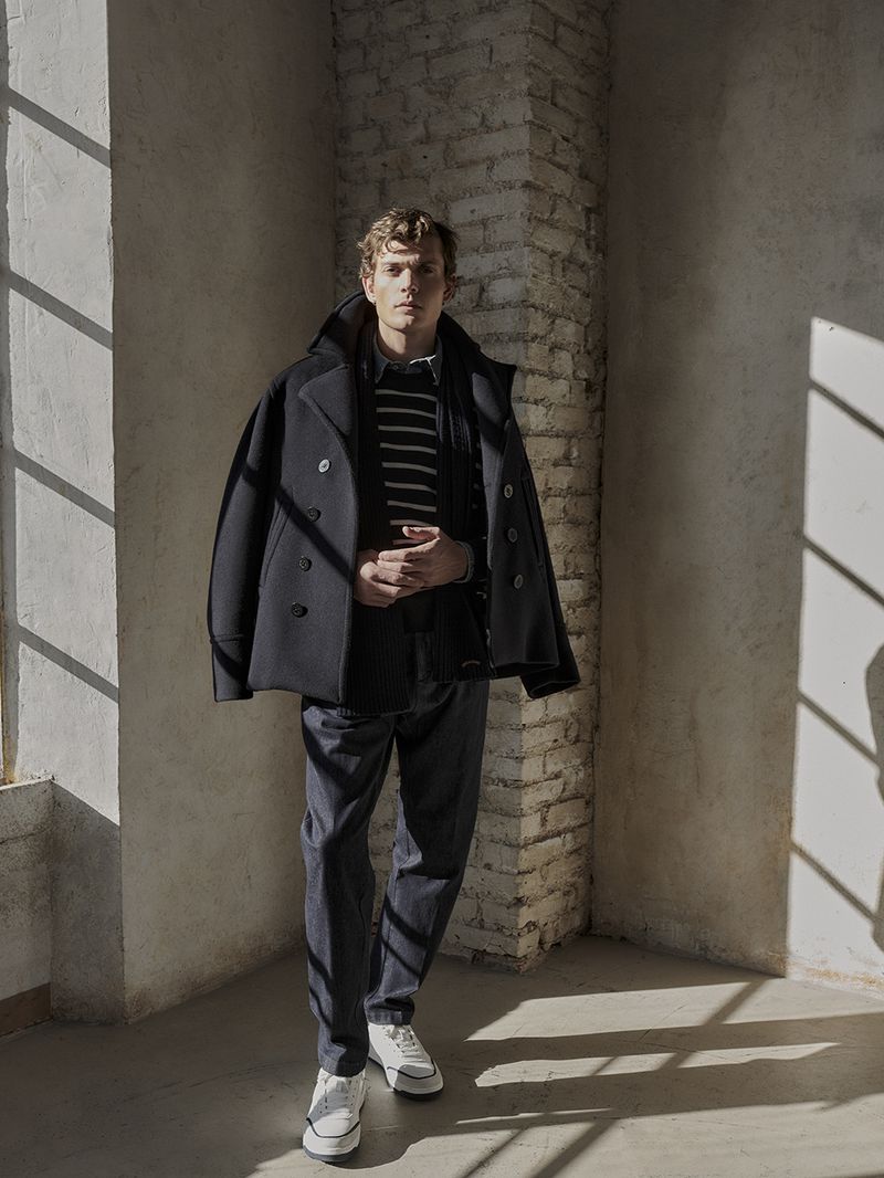 A full shot of a young man wearing a dark coat, striped sweater, and white sneakers standing in a room with light shining through a window.