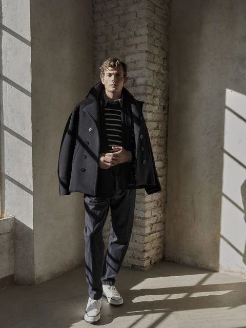 A full shot of a young man wearing a dark coat, striped sweater, and white sneakers standing in a room with light shining through a window.