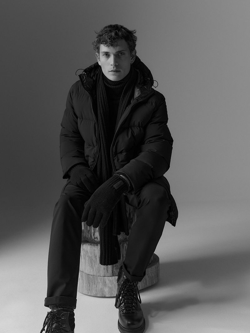 A man sits wearing a dark puffer jacket, black gloves, dark pants, and black boots on a wooden stool in a monochromatic photo studio shot.
