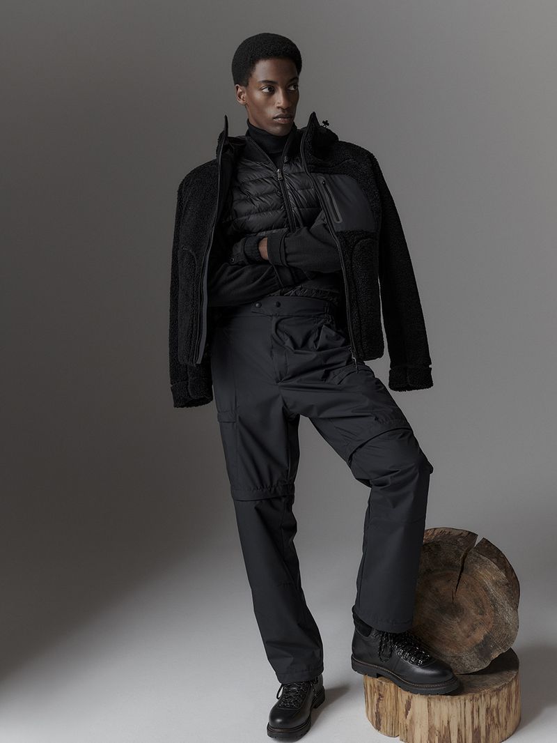 Male model poses in black outerwear and cargo pants with leather boots, standing on a tree stump against a grey backdrop in a studio.