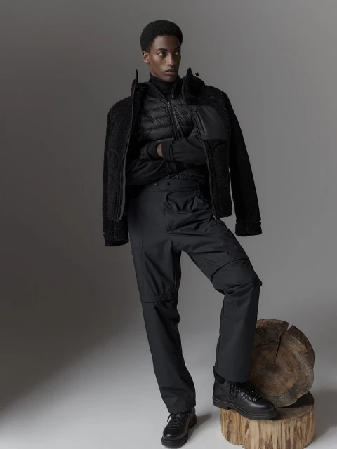 Male model poses in black outerwear and cargo pants with leather boots, standing on a tree stump against a grey backdrop in a studio.
