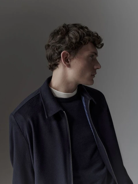 Profile of a young adult male with curly brown hair wearing a navy zip-up jacket and dark shirt against a grey gradient background.