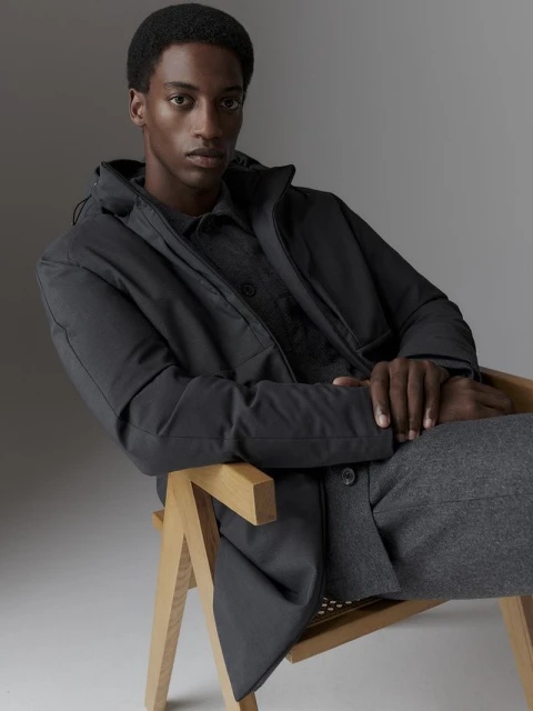An African-American man in a black jacket and grey pants sits on a light-colored wooden chair against a neutral grey backdrop in a studio shot.