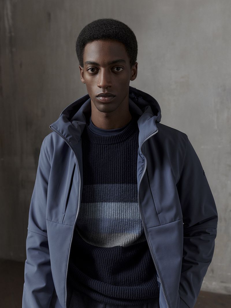 Portrait of a Black male model in a blue hooded jacket and striped sweater against a textured grey backdrop in a studio setting.