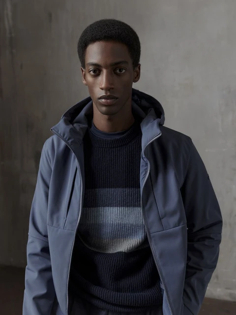 Portrait of a Black male model in a blue hooded jacket and striped sweater against a textured grey backdrop in a studio setting.
