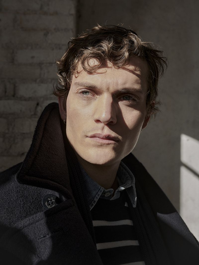 Indoor portrait of a young adult male with curly brown hair in a navy blue overcoat against a textured brick wall with natural lighting.