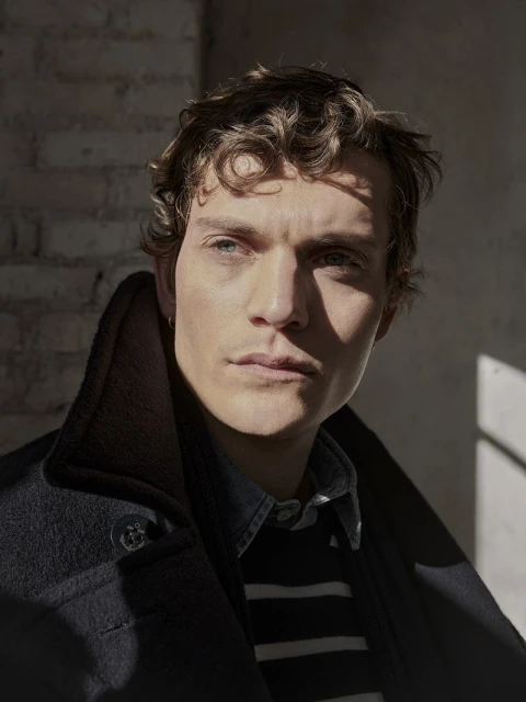 Indoor portrait of a young adult male with curly brown hair in a navy blue overcoat against a textured brick wall with natural lighting.