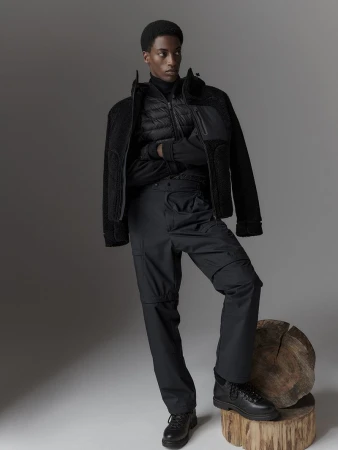 Black man in layered black winter outerwear and cargo pants posing against a neutral grey studio background with a wooden log prop.