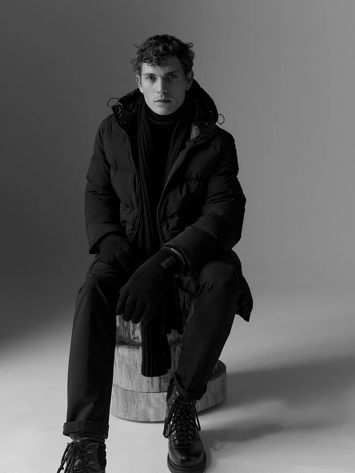 Monochrome portrait of a male model sitting on a wooden block wearing a dark puffer jacket, scarf, and boots against a gradient backdrop.