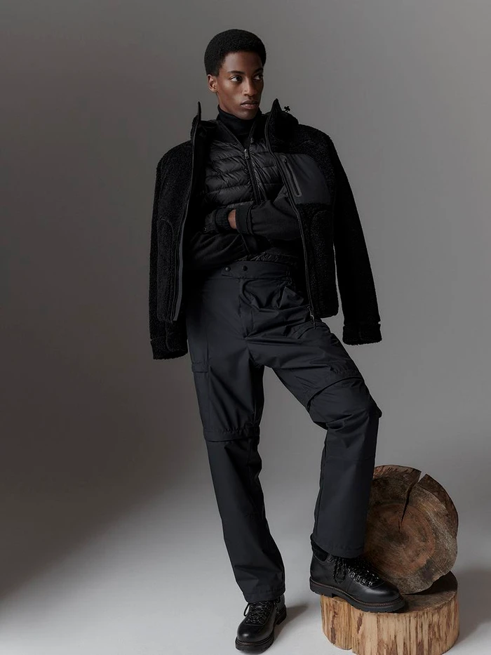Black man in layered black winter outerwear and cargo pants posing against a neutral grey studio background with a wooden log prop.