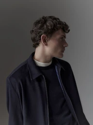 Profile portrait of a male model wearing a dark navy jacket over a white crewneck shirt against a neutral grey studio background