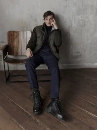Model seated on a wooden chair wearing an olive puffer jacket and dark trousers against a concrete wall