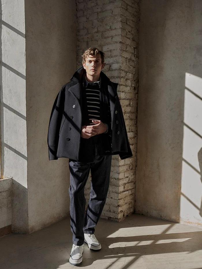 Model standing in a textured interior space wearing a dark peacoat draped over shoulders, striped sweater, and trousers.